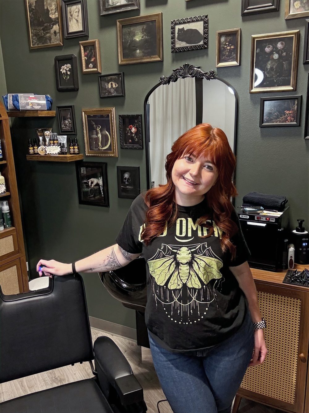 Arika, owner of Shear Madness Barbering, standing beside her barber chair in the loft, in front of a wall of framed artwork.