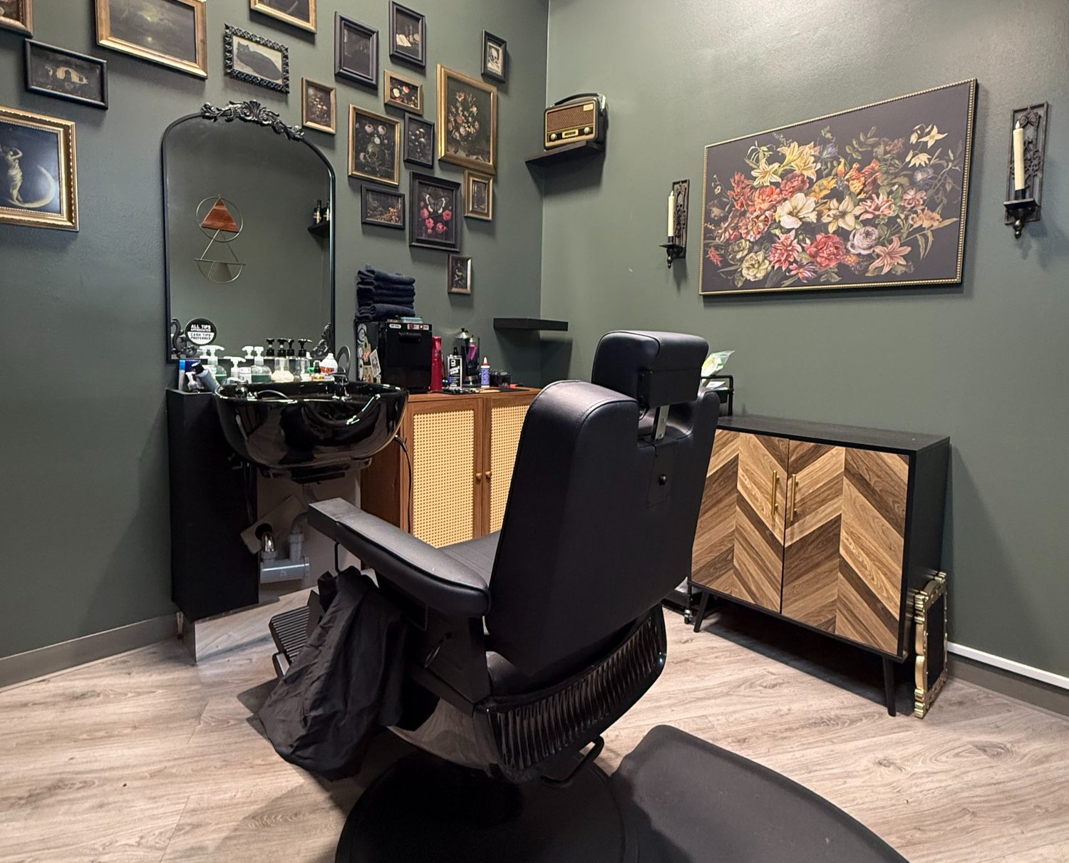 The Shear Madness loft: a black barber chair and shampoo bowl against a sage-green wall hung with framed art, beside a wood-panelled cabinet.
